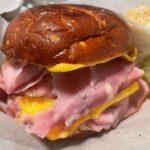 A close-up of a sandwich with layers of ham and melted cheese on a pretzel bun, served with a side of mustard and potato salad in a small plastic cup.