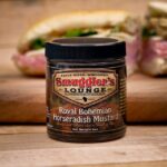 A jar of Smuggler’s Lounge Royal Bohemian Horseradish Mustard sits on a wooden surface, with a sandwich in the background.