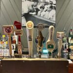 A selection of beer taps in a bar with various labels including Keweenaw, Badger, Amber, Miller Lite, Busch Light, New Glarus, and Horny Goat. A black-and-white photo hangs above.