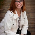 A woman with red hair and glasses, wearing a white coat, smiles while kneeling on a wooden pathway outdoors.