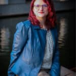 A person with shoulder-length red hair, glasses, and a blue jacket sits on a wooden bench by the water under an archway.