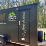 Side view of a black mobile sauna trailer from Northern Steam Co. with the slogan "Ready. Set. Sauna." and an illustration of a steamy sauna on the door. Trees and sky are visible in the background.