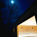 A modern sauna with illuminated interior is pictured at night under a bright moon and clear dark sky with tree silhouettes.