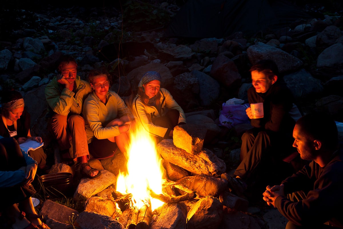 A group of people sitting around a campfire at night, smiling and holding mugs, with a tent visible in the background.