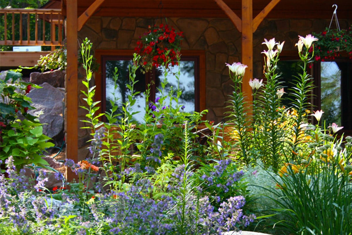 A vibrant garden with a variety of blooming flowers in front of a rustic stone house with a wooden porch and hanging plants.