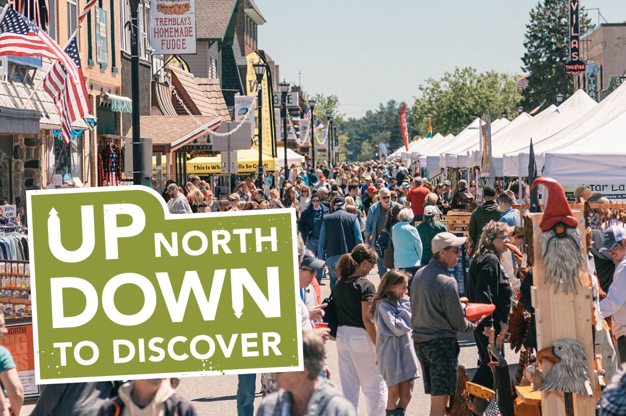 Busy street fair in a small town with vendors and shoppers on a sunny day, featuring a "up north - down to discover" logo.