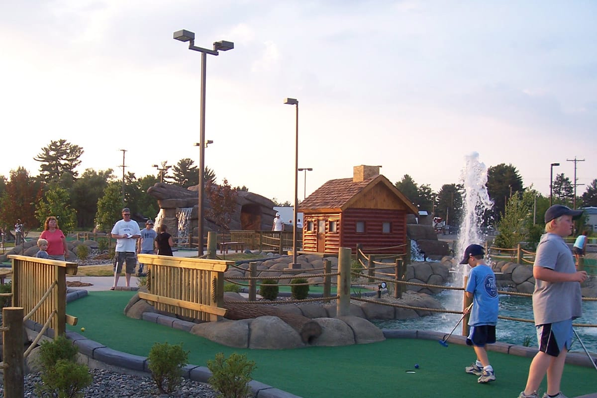 People playing mini-golf at an outdoor course with wooden structures and a water fountain under a clear sky.
