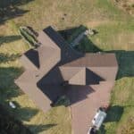 Aerial view of a large house with dark roofing, surrounded by a mowed lawn, garden beds, scattered equipment, and vehicles in the driveway.