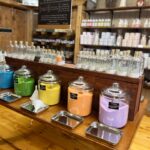 A display of various colored bath salts in large glass jars with stainless steel scoops in a rustic store setting. Shelves of bottled bath products are in the background.