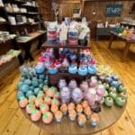 A display table in a store showcases colorful bath bombs and soaps arranged in a variety of shapes, including cupcakes. The wooden shelves in the background hold additional bath and body products.