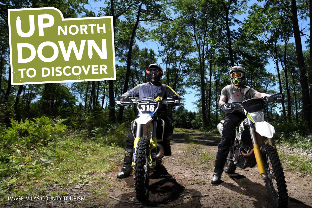 Two people on dirt bikes wearing helmets and protective gear on a forest trail, with text "up north down to discover" overlaying the image.