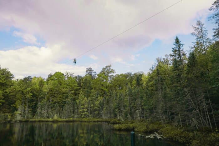 Person zip-lining over a forested lake under a cloudy sky.