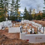 Construction site in a forested area with foundation walls being built using insulated concrete forms, surrounded by tall trees and construction equipment.