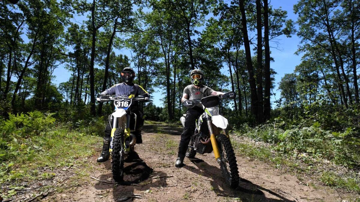 Two motorcyclists wearing full gear and helmets pause on a dirt trail in a forest, with their bikes facing the camera.