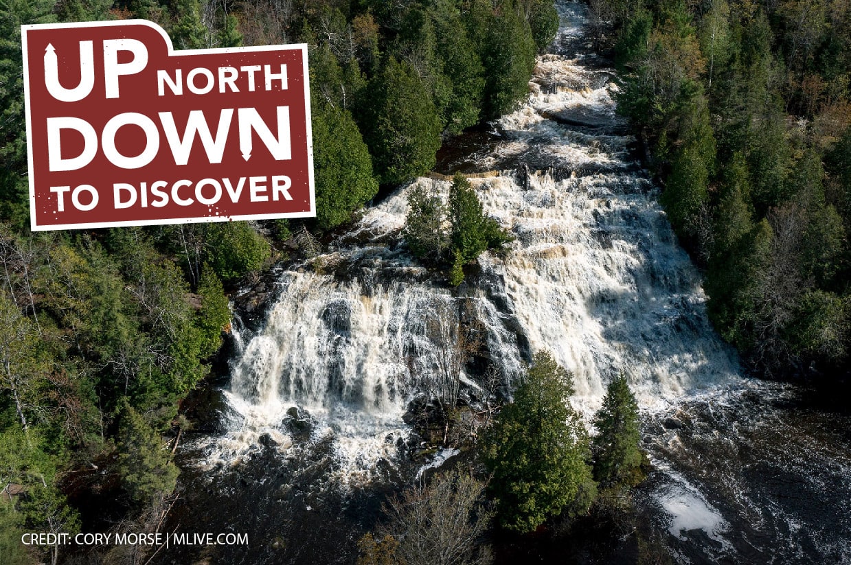 Aerial view of a cascading waterfall surrounded by dense forest. A sign in the top-left corner reads, "Up North Down to Discover". Credit: Cory Morse/MLive.com.