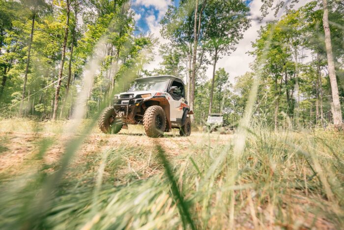 An ATV traverses a trail through a forested area with lush greenery and tall trees under a partly cloudy sky.