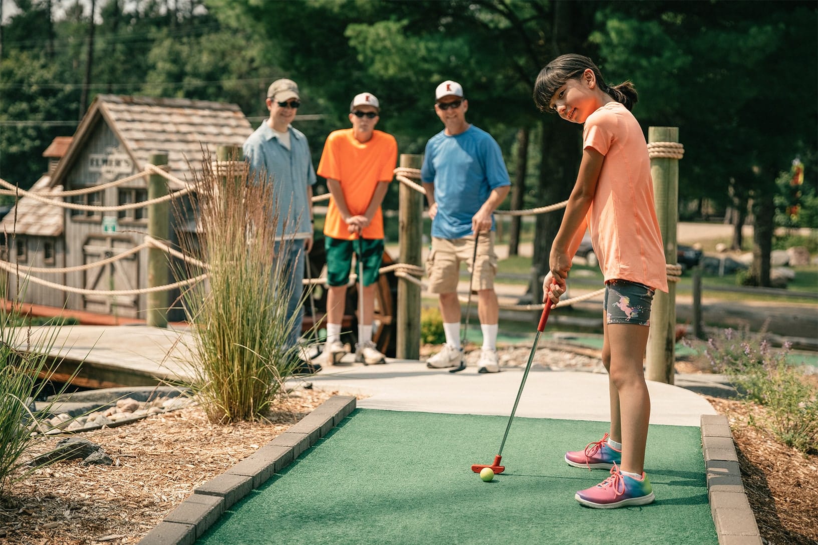 A young girl in shorts and a t-shirt plays mini-golf, preparing to putt the ball, while three people stand and watch in the background. The setting is an outdoor mini-golf course with greenery.