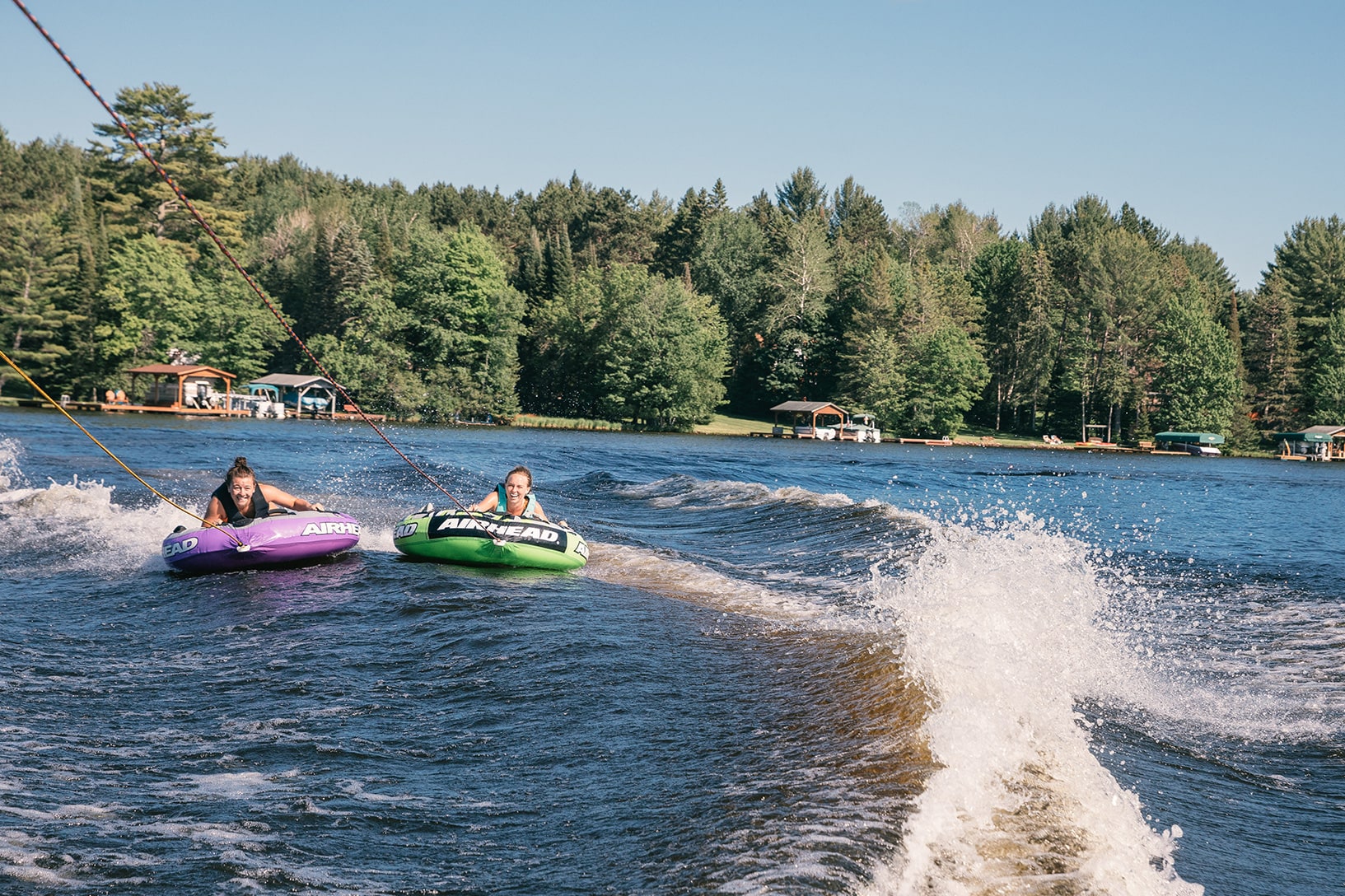 Two people are tubing on a lake, each riding in an inflatable tube pulled by a boat, creating a wake. They are surrounded by a tree-lined shore and small lakeside cabins.
