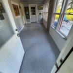 A narrow hallway with white doors on either end, gray speckled flooring, multiple windows on the right, and various framed pictures hanging on the gray walls.