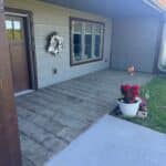 Front porch of a house with a brown door, window, potted plants with red flowers, and a white wreath hanging beside the door. The porch has a tiled floor and a small lawn area on the right.