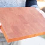 A person holding a square wooden cutting board with a smooth finish, demonstrating its craftsmanship and quality outdoors.