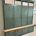 A green storage unit with four tall cabinets featuring circular cutouts, three wicker baskets on top, and four drawers at the bottom with a wooden bench in front.