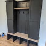 A built-in mudroom unit with black cabinets, a wooden bench, and three black hooks. Three cubby spaces are located under the bench. The floor is made of light-colored wood. Items are on the floor nearby.