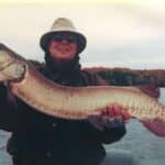 A person in a hat and sunglasses, standing outdoors near a body of water, holds a large fish horizontally with both hands.