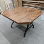 A wooden octagonal table with a natural finish and black metal legs, situated in a workshop with various wooden boards and materials in the background.