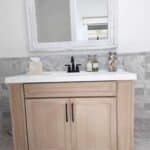 A bathroom vanity with a white countertop, a black faucet, and a large mirror above it. There are two bottles, a tissue box, and a jar of seashells on the countertop. The wall and floor have marble tiles.