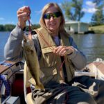 Person sitting in a boat on a lake, wearing a life jacket and sunglasses, holding up a freshly caught fish by a red string. Trees and a building are visible in the background.