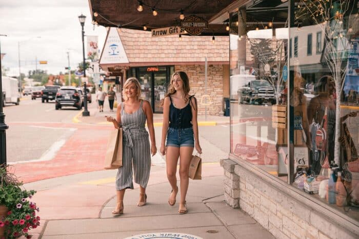 Two women walk on a sidewalk, each carrying shopping bags. They pass storefronts with displays and windows. They're spending time together for a weekend getaway up north.