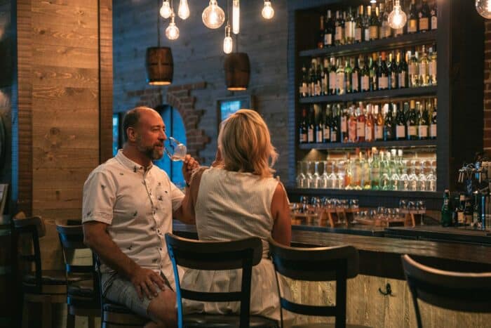 A man and a woman sit at a bar counter in a dimly lit room, drinking wine. Shelves stocked with various wine bottles are in the background, and hanging lightbulbs cast a gentle glow reminiscent of cascading waterfalls.