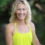 A woman with curly blonde hair is smiling, wearing a bright yellow sleeveless top, with a blurred green background.