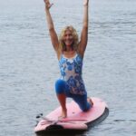 A woman kneels on a pink paddleboard with arms raised, performing yoga on a calm body of water.