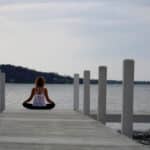 A person with light-colored hair sits cross-legged on a wooden dock facing a calm body of water, surrounded by white pillars, under a cloudy sky.