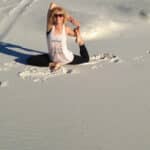 A person is practicing a yoga pose on a sandy beach. They are sitting with one leg extended and the other leg bent, touching their foot with both hands. They wear a tank top, leggings, and sunglasses.