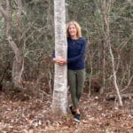 A person in a dark long-sleeve shirt and green pants stands outdoors, smiling and hugging a tree trunk in a forested area.