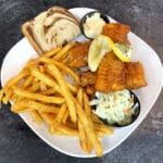 Plate with fried fish topped with lemon slices, French fries, coleslaw, a slice of bread, and a small dish of dipping sauce, perfect for pairing with your favorite brew.