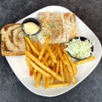 Plate with grilled fish, coleslaw, tartar sauce, fries, and a slice of marble rye bread on a dark surface, perfectly paired with a cold beer for the ultimate dining experience.