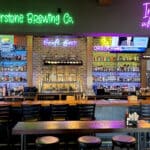 The bar interior features wooden tables, stools, and a large backlit display showcasing various liquor bottles. Neon signs glow with "Riverstone Brewing Co." and "Iron Bison whiskey bar," while TVs entice patrons with exciting beer discussions and brewing documentaries.