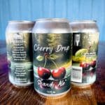 Three cans of Cherry Drop Shandy Ale, showcasing vibrant cherry graphics, rest on a wooden surface against a serene blue background—a testament to the art of brewing beer.