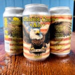 Three cans of Patriot Cream Ale, brewed with pride, showcase an eagle graphic and American flag design. The label notes 5.4% Alc/Vol and 14 IBU, embodying the spirit of American beer craftsmanship.