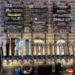 A wall-mounted draft beer setup with multiple taps and a chalkboard menu showcases a variety of brews. Each beer and cider is listed with vibrant writing and illustrations, celebrating the art of brewing.