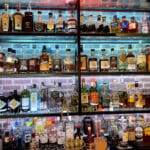 Shelves filled with various bottles of liquor, including whiskey, gin, vodka, and liqueurs, alongside a curated selection of craft beer against a backlit brick wall.