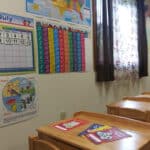 Classroom with wooden desks, educational posters, a map of the United States, and a calendar on the wall. Natural light from a window with floral curtains brightens the room.