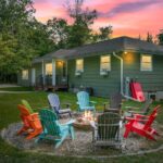 A green house with lit windows sits behind a fire pit surrounded by colorful Adirondack chairs on a grassy lawn at sunset.