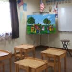 A classroom with small wooden desks and chairs, a colorful painting on an easel, an American flag, and alphabet letters displayed on the wall.