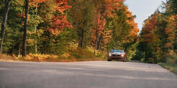 A red car drives along a rural road surrounded by trees with autumn foliage.