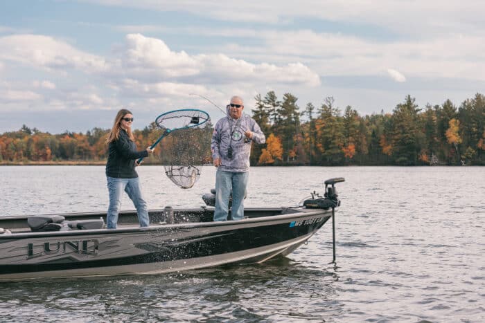 Two people on a fishing boat; one is holding a fishing net over the water while the other assists. Trees with autumn foliage and a cloudy sky are in the background.